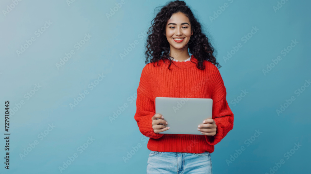 Obraz premium woman with curly hair is smiling and holding a laptop, wearing a red sweater, against a teal blue background