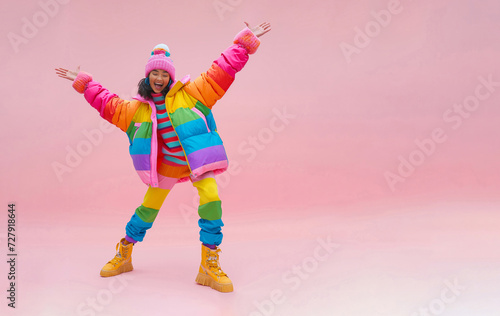Young, smiling, and happy girl dancing in front of a light pink wall in quirky, colorful clothing with rainbow colors in a winter style.