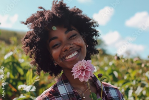 A joyful black young woman with curly hair smiles, holding a vibrant pink dahlia flower, with a sunlit field in the background.