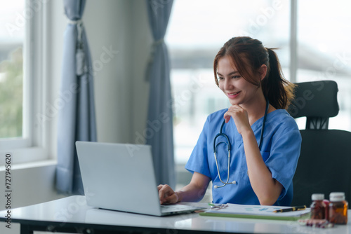 Friendly female nurse in blue scrubs seated at a desk, working on a laptop with medical charts and a stethoscope..