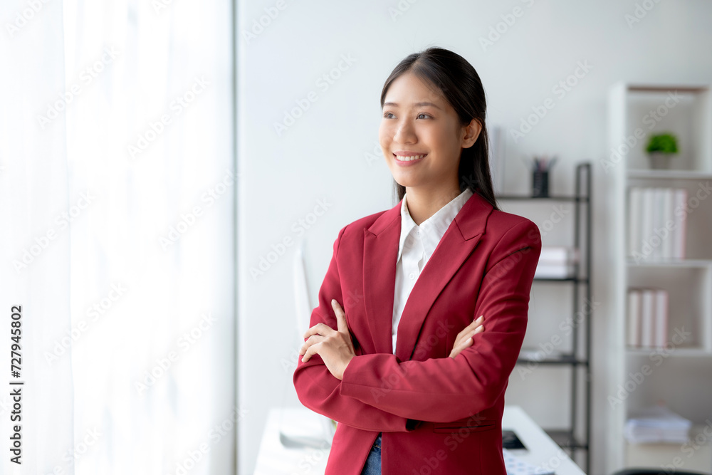 A cheerful Asian businesswoman with crossed arms smiling confidently at her clean and organized workstation in a corporate office. A cheerful Asian businesswoman with crossed arms smiling confidently at her clean and organized workstation in a corporate office.