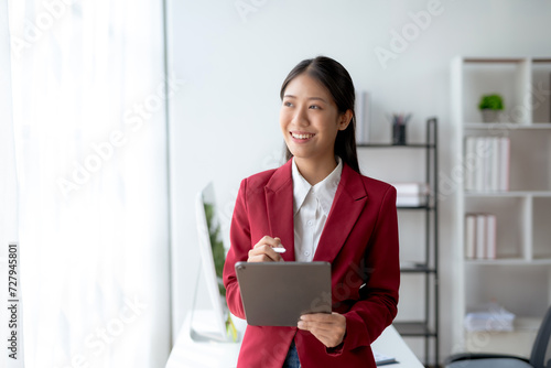 Professional young Asian businesswoman smiling while working on a digital tablet in a modern office setting.