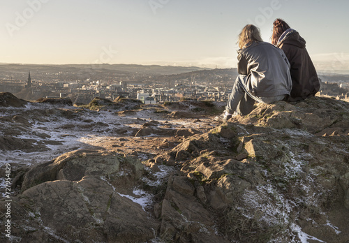 Two women sitting relaxing on top of rock over the mountain and enjoying looking at scenic view of cityscape. Two female Sitting down enjoying the amazing view from the mountain top of Salisbury Crags