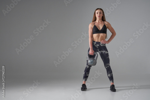 Beautiful sportswoman in activewear, lifting kettle bell in studio. Front view of strong Caucasian female with long hair holding weight, while looking at camera, copy space. Weightlifting concept.