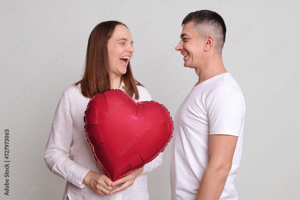 Laughing woman and man wearing white clothing holding heart shaped air balloon looking at each other expressing positive emotions satisfied couple isolated over gray background