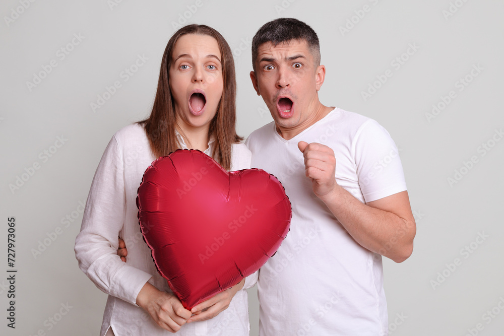 Shocked surprised astonished couple man and woman with heart shaped balloon isolated over grey background looking at camera with big eyes saying omg