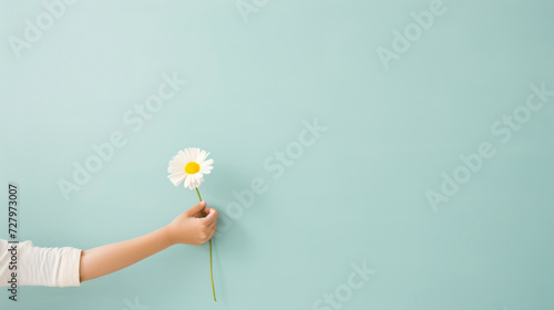 Child's hand presenting a single daisy against a soft blue background, concept of innocence and simplicity.