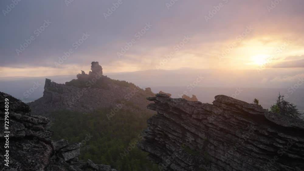 Sunlit cliffs and rock towers under a dramatic morning sky. Clouds are rapidly floating through an old mountain range overgrown with forest, piles of stones illuminated by the dawn sun Sunlit cliffs and rock towers under a dramatic morning sky. Clouds are rapidly floating through an old mountain range overgrown with forest, piles of stones illuminated by the dawn sun