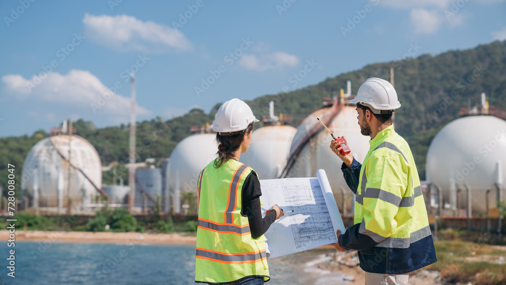 Two engineers in safety gear with blueprints discussing work in front of industrial storage tanks on a clear day. Two engineers in safety gear with blueprints discussing work in front of industrial storage tanks on a clear day.