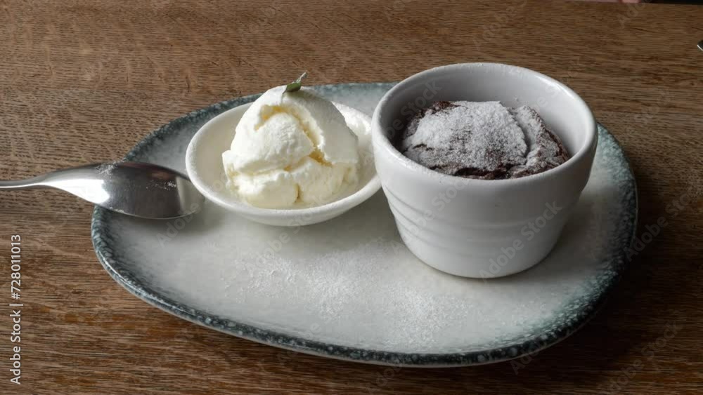 Molten chocolate lava cake and a scoop of ice cream in ceramic jars, served on a white plate with a silver spoon, on the wooden table, close-up shot. Molten chocolate lava cake and a scoop of ice cream in ceramic jars, served on a white plate with a silver spoon, on the wooden table, close-up shot.