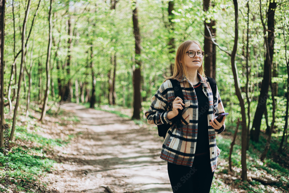 Young happy woman with backpack using smartphone and walking in the forest. Spring time. 30s Women in sport clothes hiking on woodland. Wanderlust travel concept, atmospheric moment