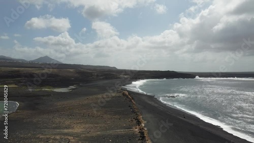 drone shot of a descent from above on black beach with clouds passing and ocean and mountains. 
lanzarote, spain