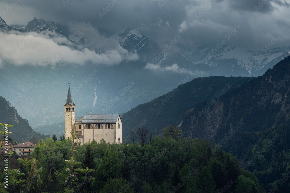 Moody landscape with small church in Dolomites mountains, Belluno, Veneto, Italy. Chiesa Parrocchiale di San Martino in Italian Alps in foggy and cloudy day at springtime. Travel destination