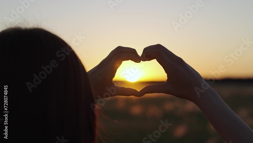 Silhouette of child of girl in park at sunset shows symbol of heart with her fingers. Concept of health, sun love. Heart shape made by fingers of teenager against background of sun. Chidhood dream sky