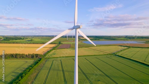 AERIAL, CLOSE UP: Spinning white windmill blades on a wind farm in rural area. Picturesque English countryside with tall modern power generation structures rising above beautifully landscaped fields. photo