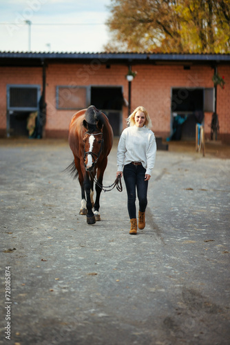 Young woman in civilian clothes leads her horse across the stables.