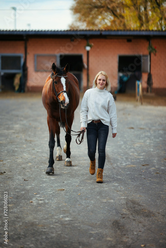 Young woman in civilian clothes leads her horse across the stables.