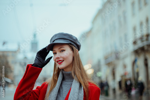 Fashionable happy smiling woman wearing elegant red coat, gray cap, black leather gloves, posing in street of European city. Outdoor close up portrait. Copy, empty, blank space for text 