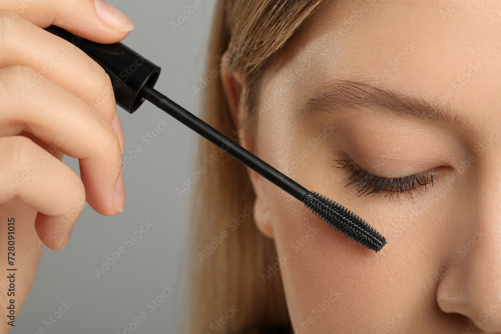 Woman applying mascara onto eyelashes against grey background, closeup