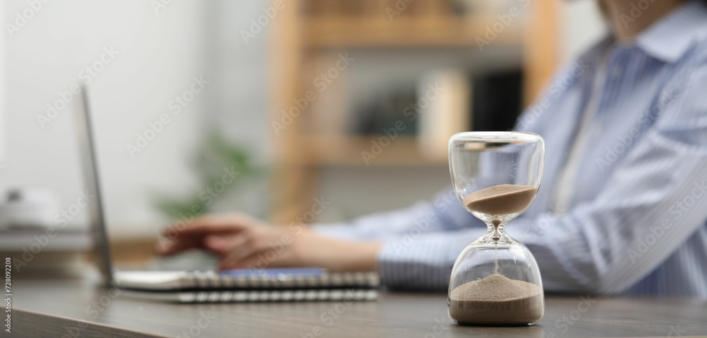 Hourglass with flowing sand on desk. Woman using laptop indoors, selective focus Hourglass with flowing sand on desk. Woman using laptop indoors, selective focus