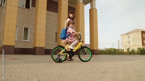 Fototapeta Naklejka Na Ścianę i Meble -  Happy family mother and little daughter riding on bike at city landmark school building together. Young caring mom teaching cute female kid child pupil with backpack driving cycle enjoy sport