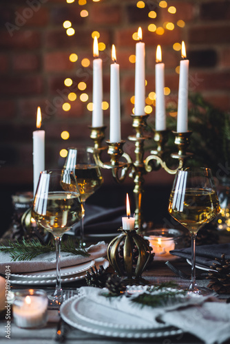 Beautiful table setting for family Christmas dinner at home. Cozy atmosphere, candlelight. Wine glasses, vintage chandelier, elegant interior. Fir tree branches, wooden furniture, dark, bokeh