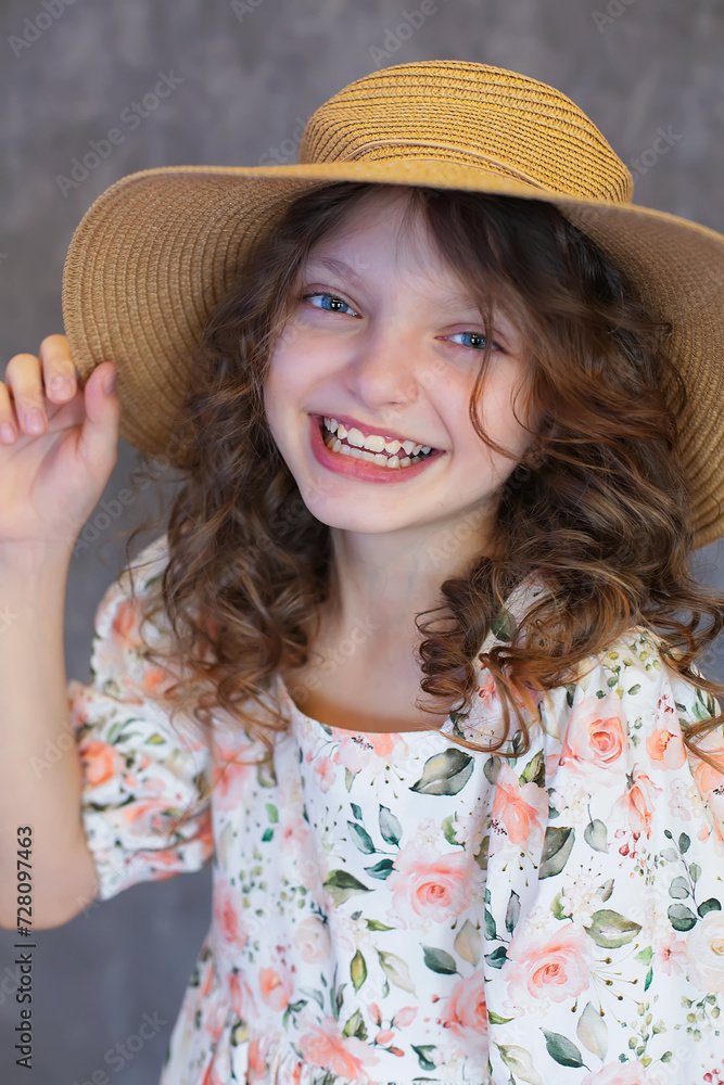 A happy teenage girl with curly hair and big blue eyes in a floral print dress and a straw hat