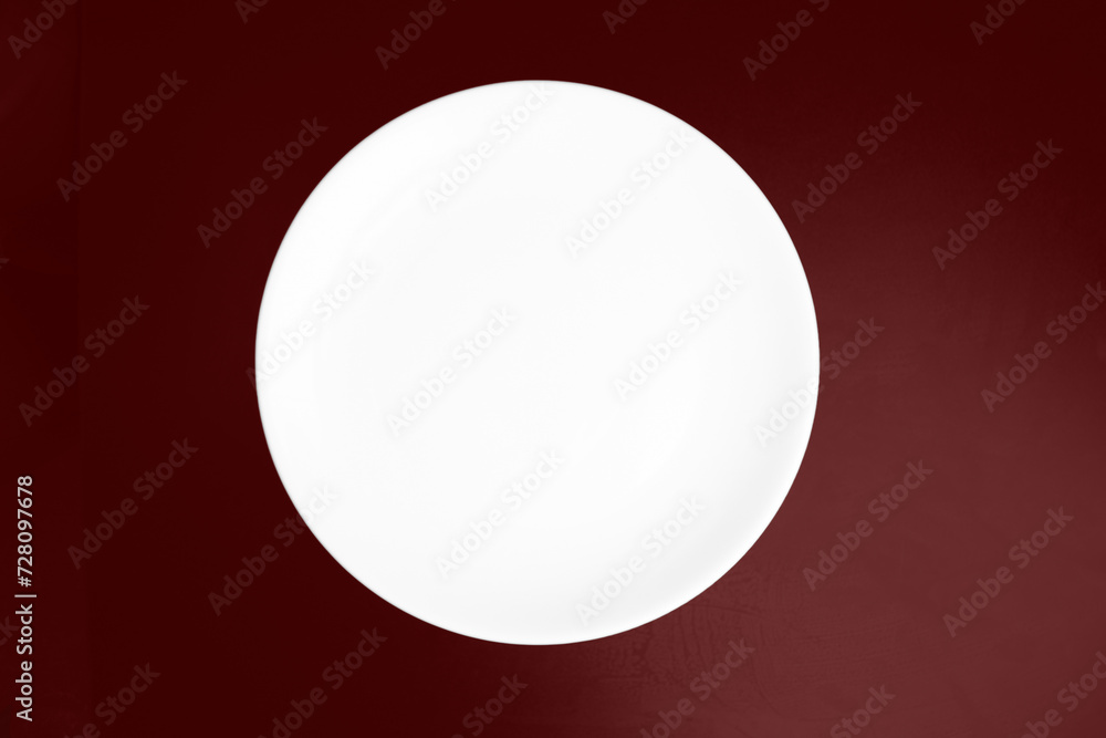White flat plate on a dark burgundy background. View from above White flat plate on a dark burgundy background. View from above