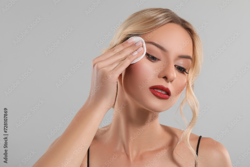 Beautiful woman removing makeup with cotton pad on light grey background