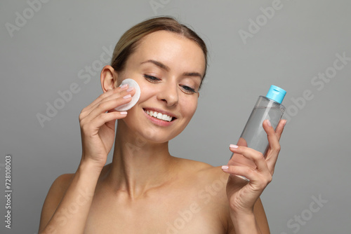 Smiling woman removing makeup with cotton pad and holding bottle on grey background