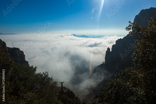 A scenic view from a high point near Barcelona, Spain, showcasing a sea of clouds flowing through mountains, with sunlight creating a lens flare and a utility pole hinting at human touch. photo