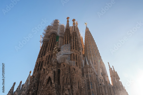 Barcelona's Sagrada Familia, by Gaudi, shines under the sun with spires touching the blue sky. Its Gothic, Art Nouveau design, active build, and Christian motifs form a stunning view.