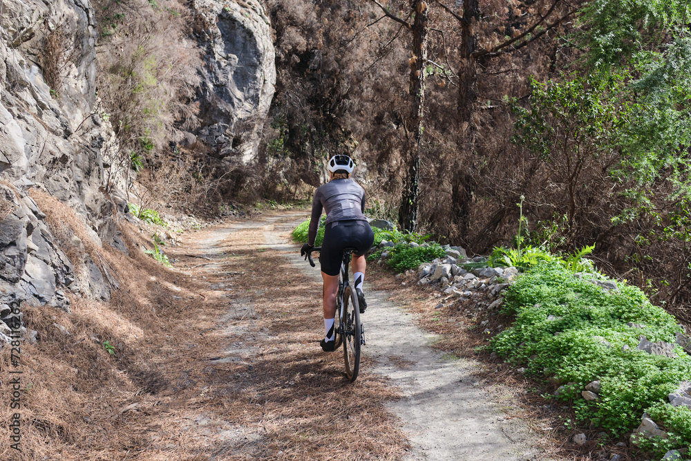 Young woman cyclist wearing cycling kit and helmet riding a gravel bike on gravel path. Biking adventure on beautiful mountain trails. Athlete riding bicycle in the countryside during vacation. Spain