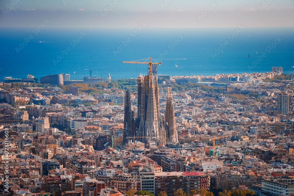 Panoramic view of Barcelona featuring the unfinished Sagrada Familia with its spires and cranes, amidst a grid of streets and diverse architecture, with the Mediterranean Sea in the backdrop.