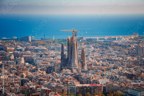 Panoramic view of Barcelona featuring the unfinished Sagrada Familia with its spires and cranes  amidst a grid of streets and diverse architecture  with the Mediterranean Sea in the backdrop.