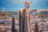 Closeup view of the Sagrada Familia's spires in Barcelona, Spain, with cranes indicating ongoing work, the cityscape, and the Mediterranean Sea in the background, bathed in warm afternoon light.