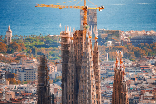 Closeup view of the Sagrada Familia's spires in Barcelona, Spain, with cranes indicating ongoing work, the cityscape, and the Mediterranean Sea in the background, bathed in warm afternoon light. photo