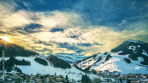 A picturesque snowy landscape captures a serene village nestled between majestic snow-capped mountains under a dramatic sky at sunset, epitomizing the concept of winter wonderland and peaceful retreat