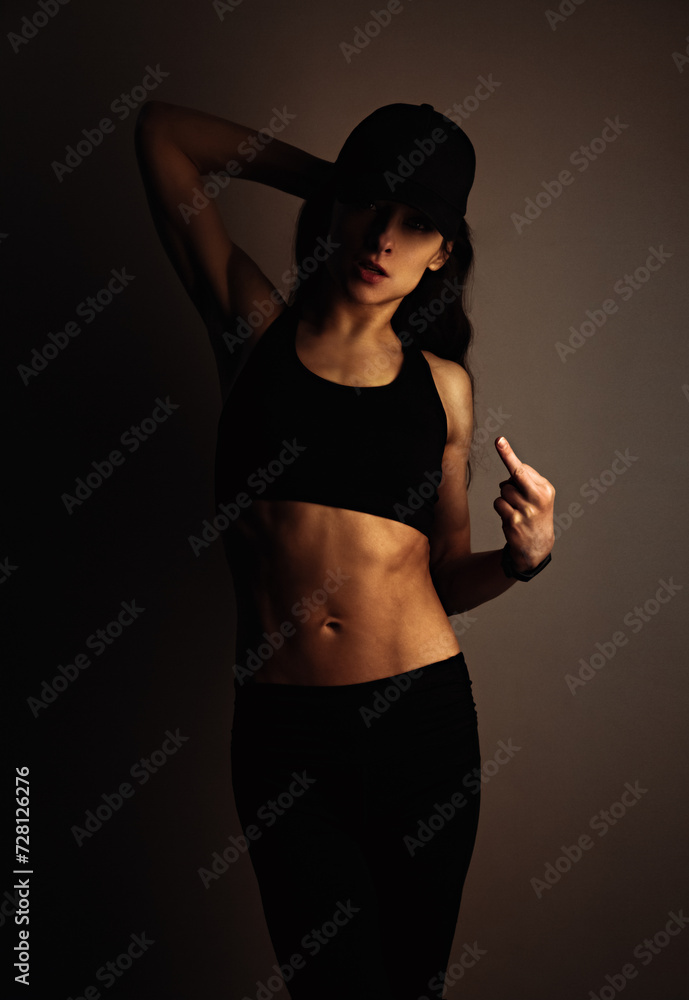 Fuck you. Sport sexy muscular woman posing in black sport bra, cap and showing the fuck sign the hand, standing on dark shadow studio background. Front body view. Fuck you. Sport sexy muscular woman posing in black sport bra, cap and showing the fuck sign the hand, standing on dark shadow studio background. Front body view.