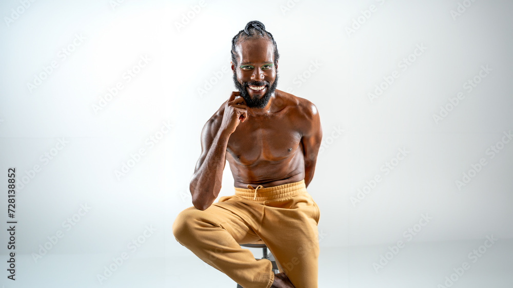 Portrait of smiling African American gay man with bright makeup. Shirtless muscular man sitting on stool and looking at camera. LGBTQ person is posing on white background. Portrait of smiling African American gay man with bright makeup. Shirtless muscular man sitting on stool and looking at camera. LGBTQ person is posing on white background.