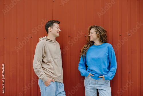 A couple of friends, a boy and a girl, are talking on the background of a red wall outdoors.