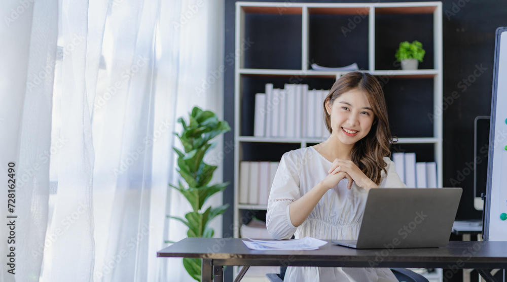 Cheerful Asian woman working with laptop in office, happy in formal suit working in office Charming smiling female office worker, financial accounting concept Cheerful Asian woman working with laptop in office, happy in formal suit working in office Charming smiling female office worker, financial accounting concept