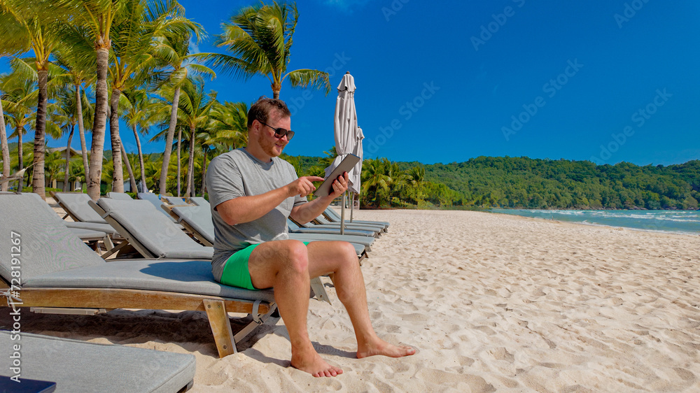 Man in casual summer clothing sitting on a beach chair and reading a tablet on a sunny tropical beach with palm trees, symbolizing leisure and vacation
