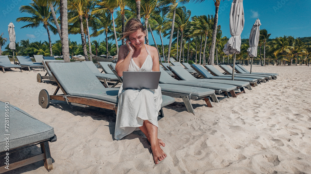 Young woman in a white dress working on a laptop while sitting on a sunbed at a tropical beach resort with palm trees