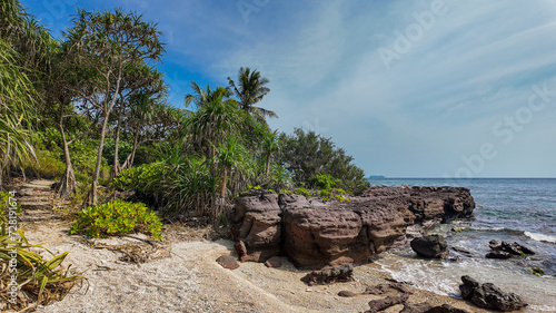 Sunny tropical beach with rocky shoreline and lush greenery under a vast blue sky, evoking a serene tropical vacation concept