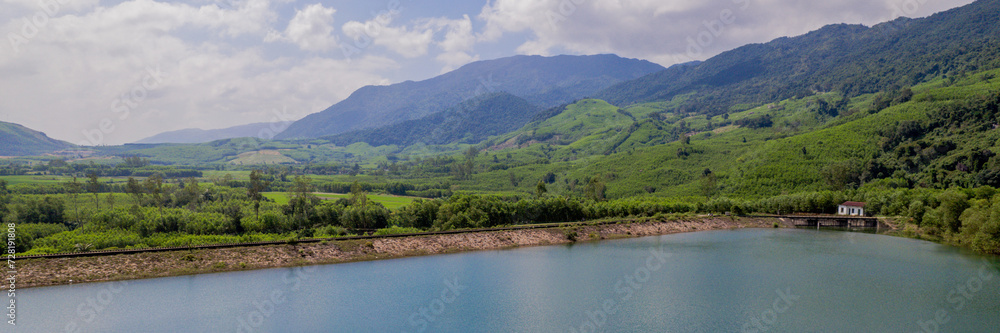 A panoramic view of a tranquil reservoir nestled in a lush valley with green mountains in the background, under a hazy sky