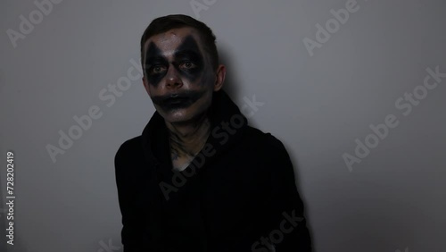 A man with a black clown makeup mask sits down at the floor and stares at the camera photo