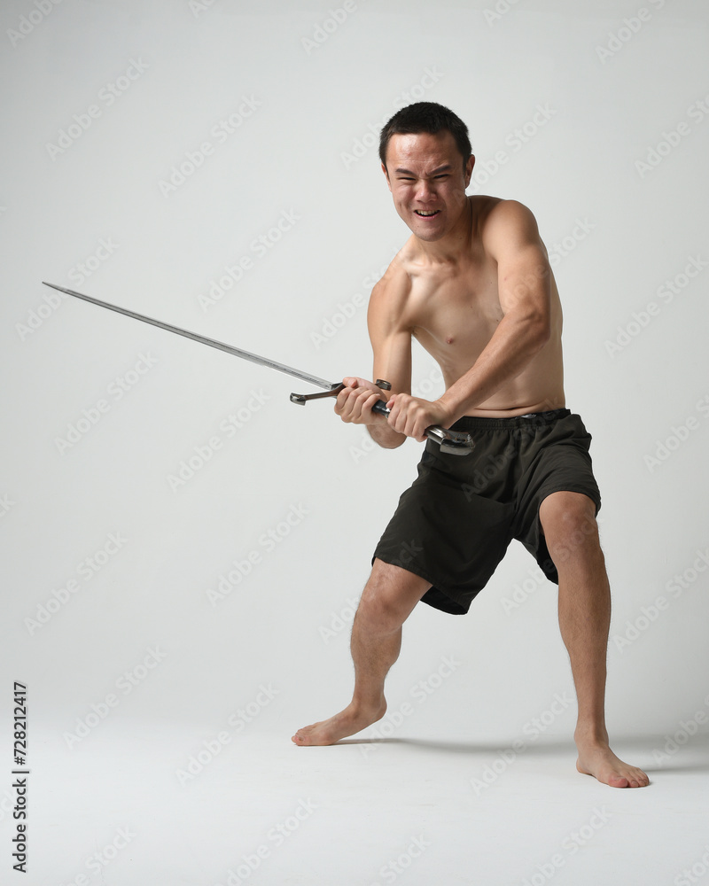 Full length portrait of fit handsome asian male model, wearing gym shorts and shirtless. Holding sword weapons whilst in warrior training action poses, isolated on white studio background. Full length portrait of fit handsome asian male model, wearing gym shorts and shirtless. Holding sword weapons whilst in warrior training action poses, isolated on white studio background.