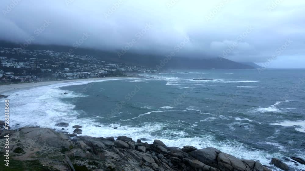Moody aerial view of camps bay in Cape Town with rough sea and waves crashing on rocks during a winters day Moody aerial view of camps bay in Cape Town with rough sea and waves crashing on rocks during a winters day