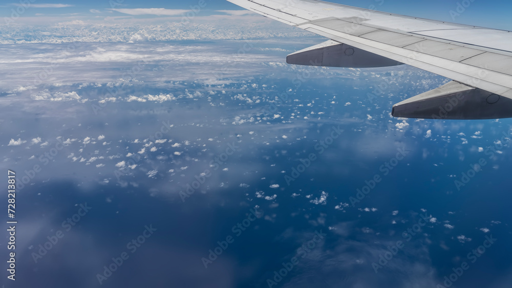 Ocean view from the plane. White clouds are scattered over the endless blue expanse of water. Reflection in the sea. In the foreground is an airplane wing.